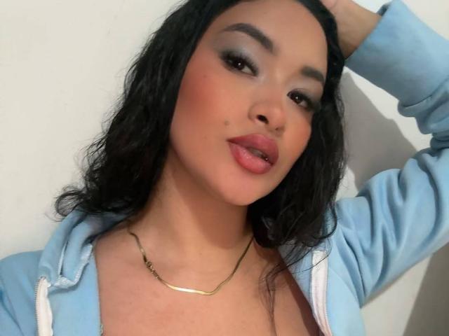AdalinaVera with wavy black hair, light blue hoodie, gold necklace, and pink lipstick, holding her head with a slight smile, showing a small beauty mark on her cheek.