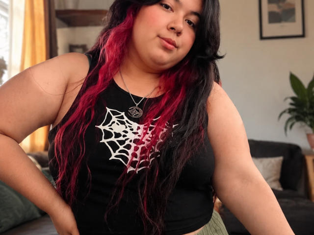 SultanaLm, une femme corpulente avec de longs cheveux noirs et roux, porte un débardeur noir en forme de toile d'araignée, debout, les mains sur les hanches, et regarde légèrement vers le bas..