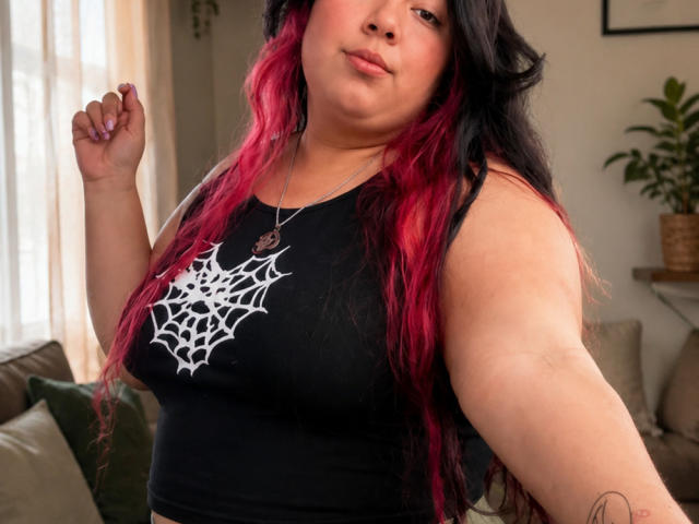 SultanaLm, avec de longs cheveux noirs et roux, porte un débardeur noir laissant apparaître un motif d'araignée blanche, et a un petit tatouage sur le bras droit. Elle se tient debout, une main levée, et regarde l'appareil photo..