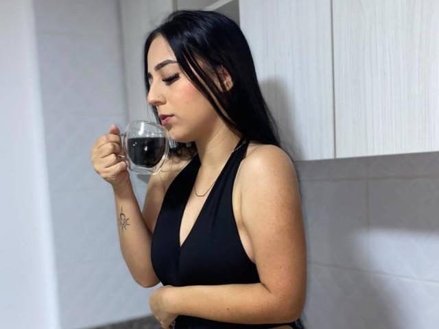 StefanyDelly with black hair, wearing a black halter top, holding a glass of red wine, looking down, small tattoo on her right wrist, white cabinets in the background.
