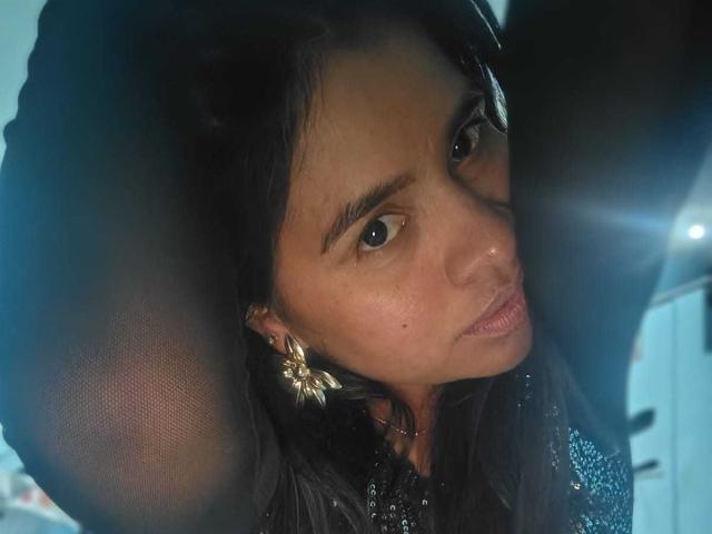 EmilyVidal with dark hair, wearing a sheer black top, exposing her tits, looking at the camera with a slightly pouty expression, a flower earring in her right ear.