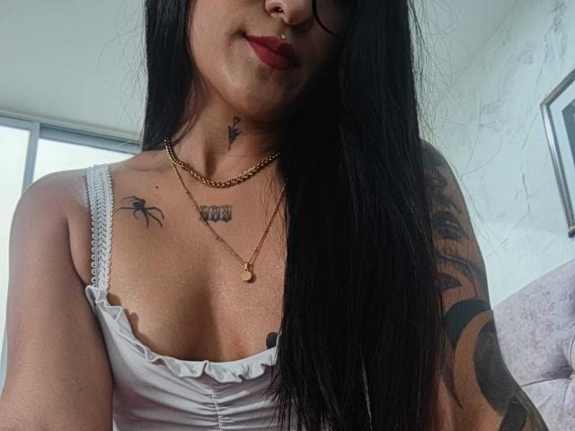 LuciadelaVega with long black hair, wearing a white tank top, showing small tits, tattoos on chest and arm, nose ring, and red lipstick, and a gold chain necklace.