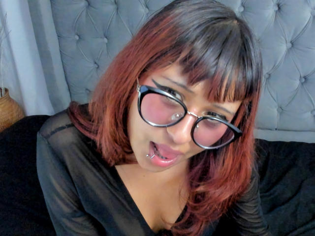 AnyOlsen, con cabello castaño rojizo, gafas negras y un piercing en el labio, vestida con una blusa negra transparente, con la boca abierta, mirando hacia arriba, con una cama negra de fondo..