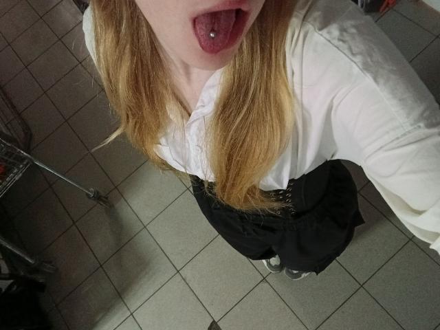 NievesLamanna with long blonde hair, white shirt, black skirt, and tongue out, showing a tongue piercing. She's looking down at the camera.