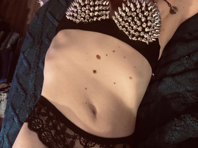 TifolyLaurent's bare midriff with small dark moles, wearing a black bra with silver spikes and black lace panties, lying on a green textured blanket.