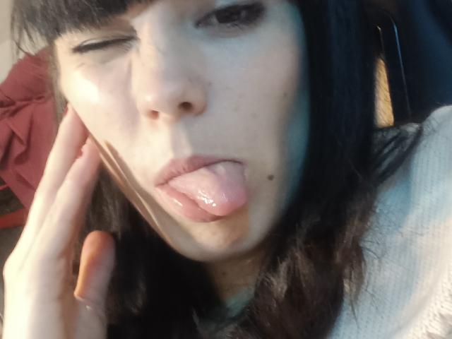 OrianaLaFrancaise's face, winking with tongue out, dark hair, pale skin, slight blush, hand near cheek, wet tongue, small mole above lip, wearing white top.