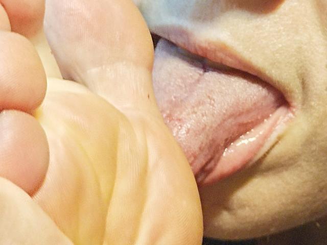 La lingua di OrianaLaFrancaise sta leccando un piede. La sua bocca è aperta, la lingua avvolge la pianta del piede, con i denti leggermente visibili. Le sue labbra sono divaricate, mostrando la sua lingua umida..