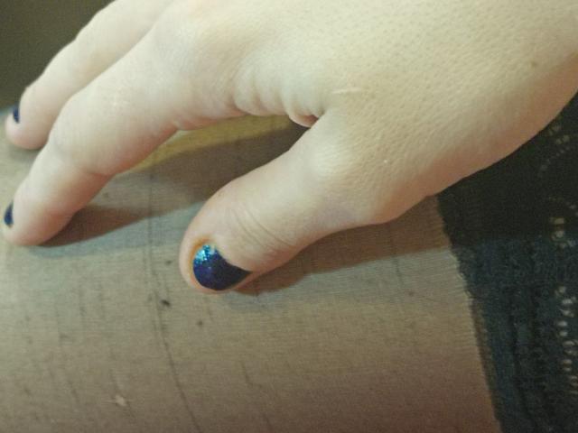 La mano di OrianaLaFrancaise, con lo smalto per unghie blu scintillante sull'indice, appoggiata su una superficie di legno. La sua pelle è chiara e le sue unghie sono corte e curate..