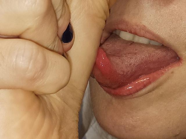 La lingua di OrianaLaFrancaise è fuori, premuta contro le sue labbra rosse, con una mano che le stringe il labbro superiore. La sua lingua è leggermente umida e lucida. Le sue unghie sono dipinte di nero..