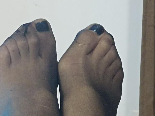 Os pés de OrianaLaFrancaise estão calçados com meias pretas finas, com os dedos visíveis e unhas dos pés azul-escuras, mostrando um leve desgaste e rugas na meia..