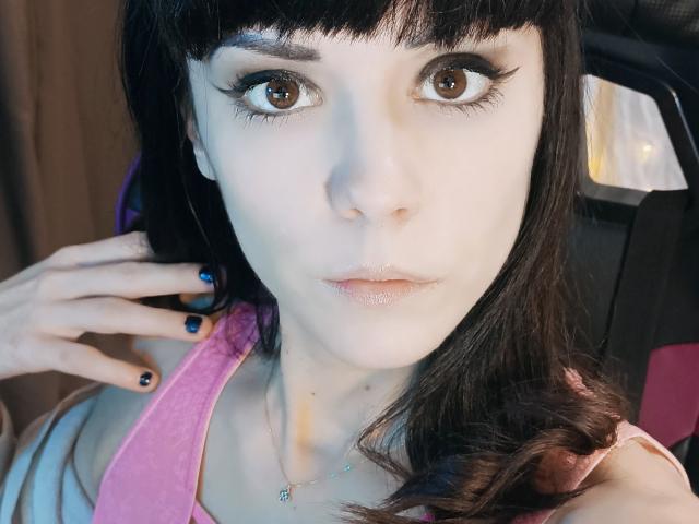 OrianaLaFrancaise has pale skin, brown eyes, and black eyeliner. She's wearing a pink top, has black nails, and a necklace with a small pendant. Her face is slightly shiny.