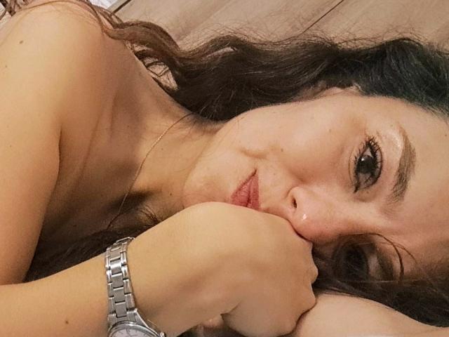 Monica Belluci está deitada em uma cama, com a cabeça apoiada na mão, cabelos castanhos ondulados, olhos escuros e um sorriso discreto, usando um relógio de prata, sem blusa, mostrando seus seios..