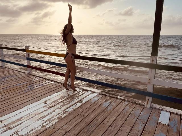 MonicaBellox, in un bikini marrone, è in piedi su un molo di legno consunto, con le braccia alzate, rivolta verso l'oceano. Ha i capelli ricci e un sedere slanciato e tonico..