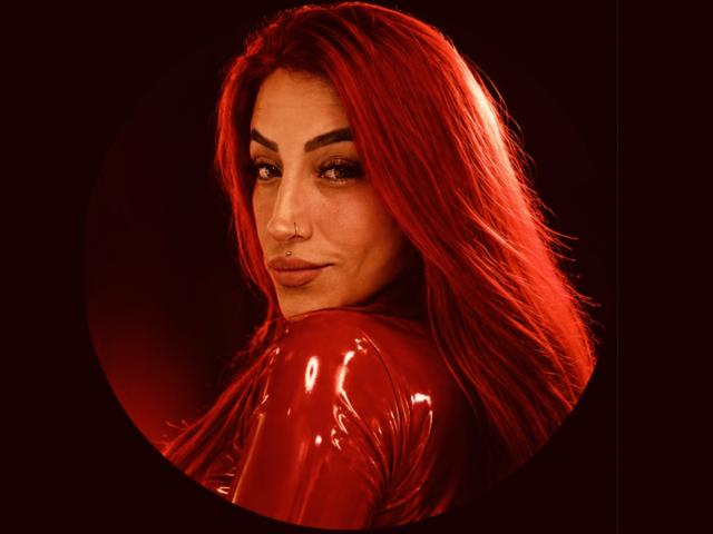 Alissa Brown, avec un haut en latex rouge et brillant, regardant par-dessus son épaule, montrant son anneau de nez, et avec ses cheveux roux, dans un cadre circulaire, dégage une aura mignonne, sexy et confiante..