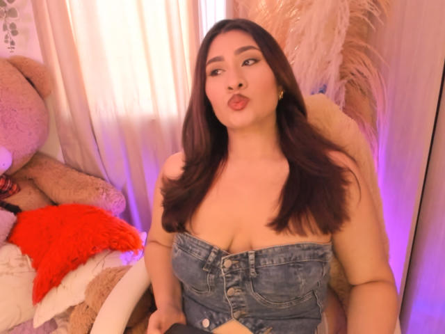 ElyRoss with dark brown hair, pouty lips, wearing a strapless denim top, exposing her tits, sitting in a chair with a red pillow and large teddy bear.