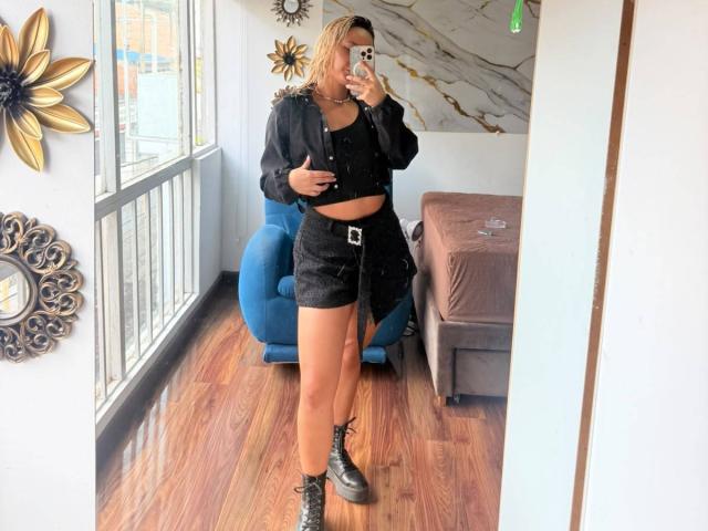 Elena Saenz vestindo uma cropped preta, shorts pretos com cinto, botas pretas, segurando um celular, selfie no espelho, cabelo loiro, em pé em um chão de madeira, uma cadeira azul, uma cama marrom, uma parede de mármore..
