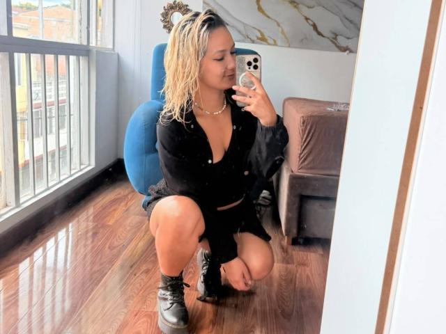 ElenaSaenz agachada em frente a um espelho, com o cabelo loiro molhado, camisa preta de botões, shorts pretos, botas pretas, segurando um celular, tirando uma selfie, mostrando os seios..