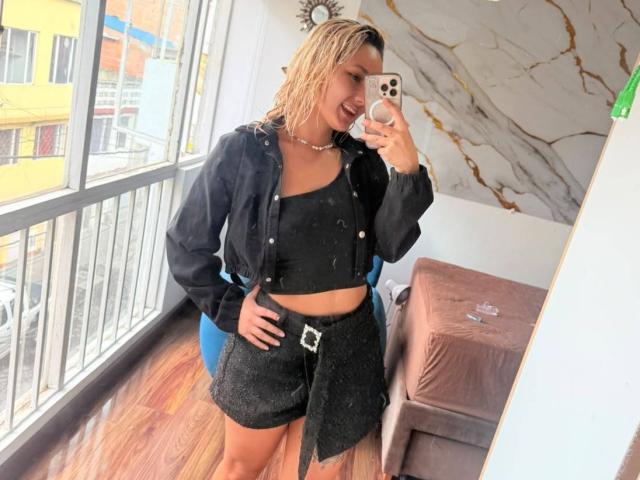 Elena Saenz, com o cabelo loiro molhado, vestindo uma cropped preta, jaqueta preta e shorts pretos com um cinto, tirando uma selfie no espelho, mostrando seu traseiro..