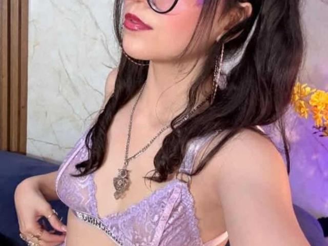 ValuandDani mit Brille, lila Spitzen-BH, mittelgroße Brüste, welliges schwarzes Haar, roter Lippenstift, Kette, goldene Ohrringe, Hände an den Hüften, leichtes Lächeln, weiches lila Licht..