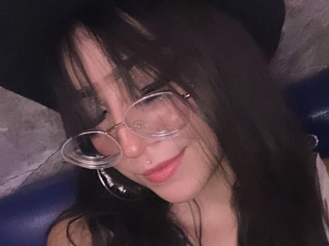 ValuandDani, con capelli neri, occhiali e un piercing al naso, sorride, indossa un cappello nero e rossetto rosa, e guarda verso la telecamera. Il suo seno è parzialmente visibile..