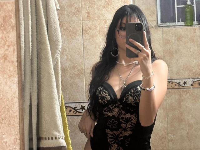 ValuandDani with long black hair, glasses, and hoop earrings, taking a mirror selfie in a bathroom. She's wearing a black lace corset, showing her boobs, and multiple necklaces.