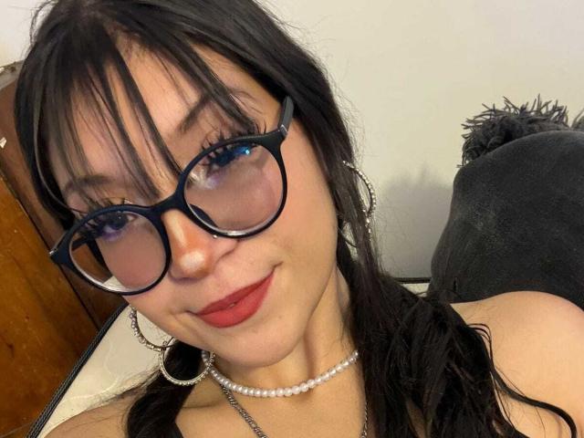 ValuandDani with black glasses, red lipstick, pearl necklace, and hoop earrings, smiling at the camera, showing her face and upper chest. She has dark hair and blue eyeshadow.