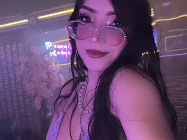 ValuandDani, com cabelos longos e pretos, usando óculos rosa, batom roxo e um colar de contas, sorrindo para a câmera, mostrando os seios em uma blusa com decote baixo..