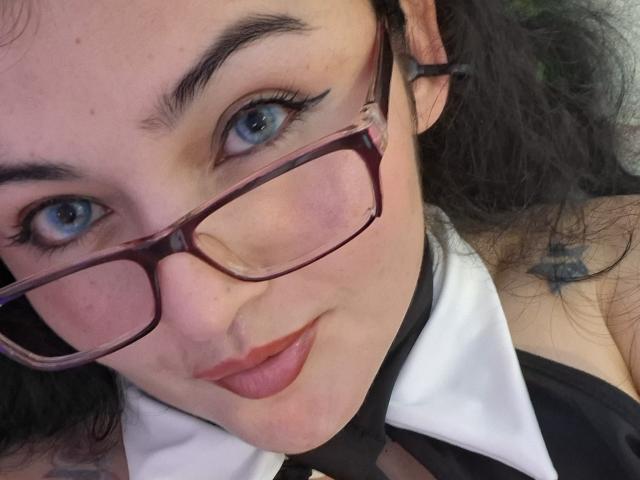 LindaSophieX met blauwe ogen, zwart krullend haar en rode lippenstift, draagt een bril en een zwart-witte kraag, en toont een kleine sterretje tatoeage op haar rechter schouder..