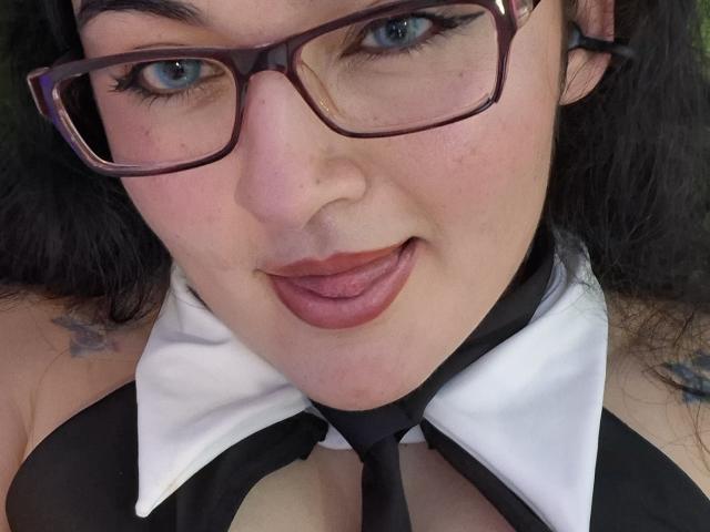 LindaSophieX, avec des lunettes, la langue tirée, portant un col noir et blanc, dévoilant sa poitrine, avec des tatouages sur les épaules, des cheveux foncés et du rouge à lèvres, dans un autoportrait..