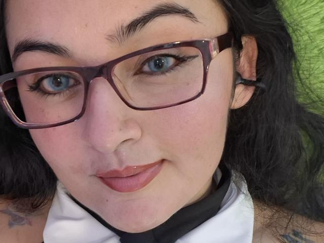 LindaSophieX with blue eyes, dark glasses, and pink lipstick, lying down, showing a slight smile, her black hair and tattoo partially visible. Her face is centered in the shot.