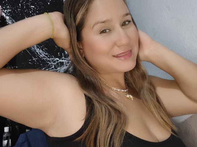 AlexaHotSeins con cabello castaño, sonriendo, con los brazos levantados, vistiendo una camiseta negra, un collar y una pulsera verde, mostrando sus pechos y un rostro atractivo, de pie en una habitación..