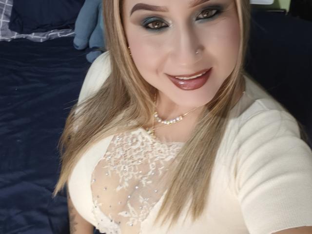 AlexaHotSeins con capelli biondi, trucco occhi blu, piercing al naso, top di pizzo bianco che mette in mostra il suo seno prosperoso, sorridente, che indossa una collana di perle, su un letto blu scuro..