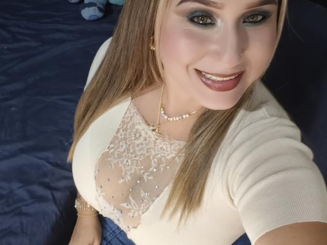 AlexaHotSeins con cabello rubio largo, sonriendo, vestida con una blusa blanca de encaje transparente, mostrando sus grandes senos, joyas doradas y maquillaje oscuro, sentada en una cama azul..