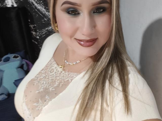 AlexaHotSeins ha i capelli biondi, l'ombretto blu e il rossetto rosa lucido, indossa una camicetta bianca in pizzo che mette in mostra il seno, e una collana di perle, sorridendo con un piercing al naso..