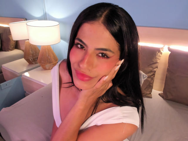 EvaGate with black hair, pink lips, white top, hand on cheek, smiling, brown eyes, soft lighting, bed with brown pillows, two white lamps with brown bases, bedroom.