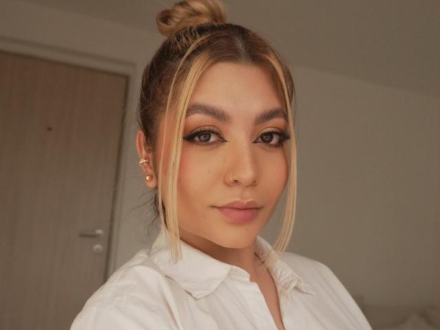 LauraDeluxe heeft blond haar in een knot, bruine ogen met zwarte eyeliner, een neusring, en draagt een witte blouse. Ze ziet er schattig uit met een lichte glimlach..