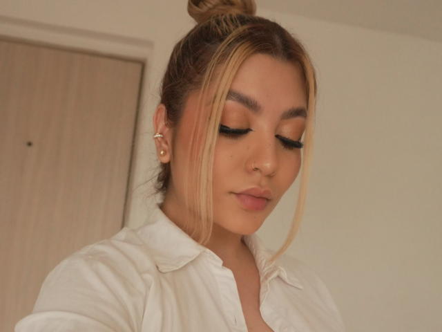 LauraDeluxe, avec ses cheveux blonds attachés en chignon, portant une chemise blanche, les yeux fermés, un trait d'eye-liner noir et un anneau de nez, regarde vers le bas avec un léger sourire..