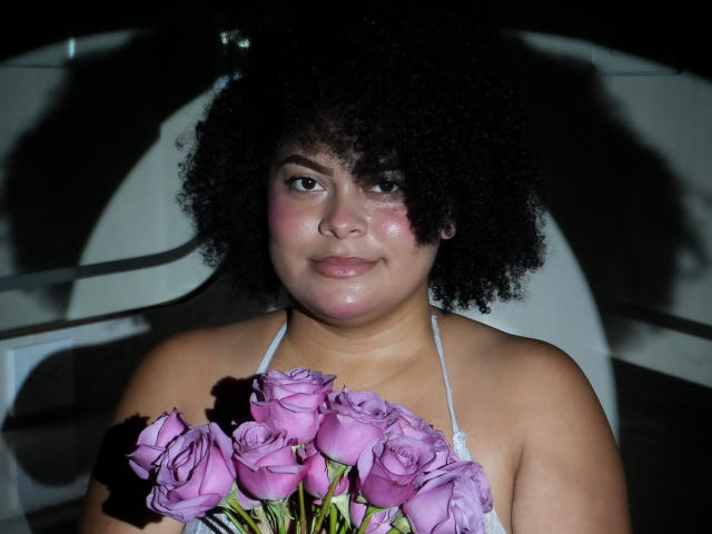 ElizabethBloom with curly black hair, holding a bouquet of pink roses, wearing a white halter top, looking slightly to the side, with a small red mark on her cheek.