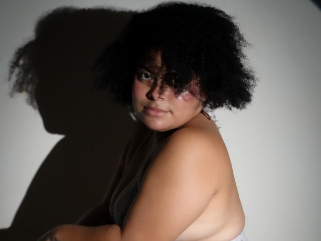 ElizabethBloom with curly black hair, looking at the camera, has a bruise on her right cheek, wearing a gray top, shadow on the wall, and a confident expression.