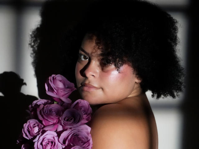 ElizabethBloom with curly black afro, holding purple roses, looking back with shiny pinkish skin, no clothes, soft light, shadow on right, serious expression, subtle makeup.