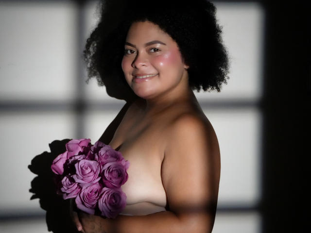 ElizabethBloom, with dark curly hair, smiling, holding a purple rose bouquet, covering her bare tits with the flowers, has a cute, chubby, brown-skinned face.
