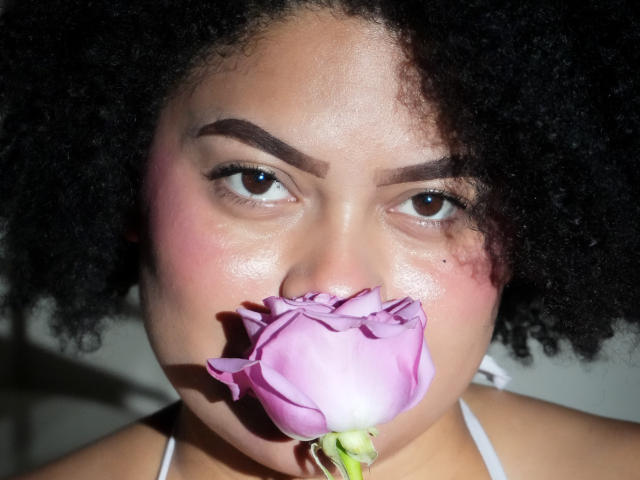 ElizabethBloom with dark curly hair, holding a pink rose between her puffy, shiny, pinkish-tan lips. She has thick, arched eyebrows and brown eyes.