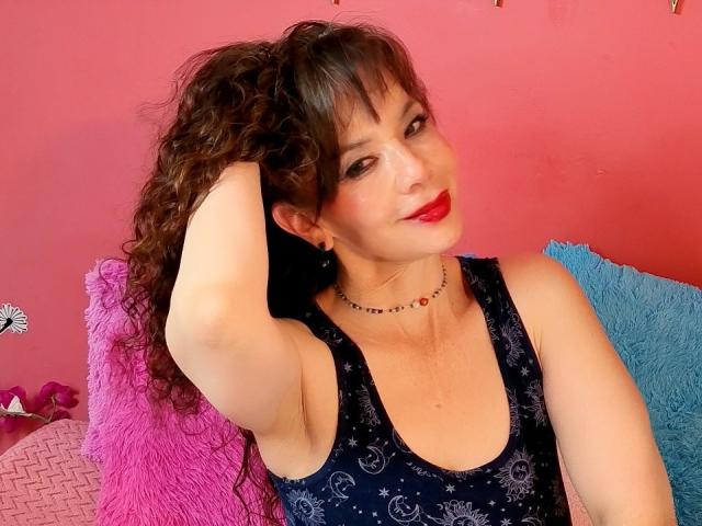 SamaraBellet, com cabelos castanhos cacheados, batom vermelho, vestindo uma regata escura com padrão azul, levantando o braço direito, sentada em almofadas rosa e azuis. Ela está sorrindo levemente..