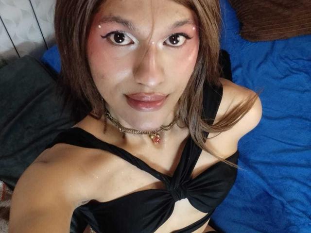 PamelaXFox mit braunen Haaren, pinkem Augen-Make-up und einem schwarzen Top ohne Träger, das kleine Brüste zeigt, eine Halskette und ein süßes Gesicht, sitzend auf einem blauen Bett..