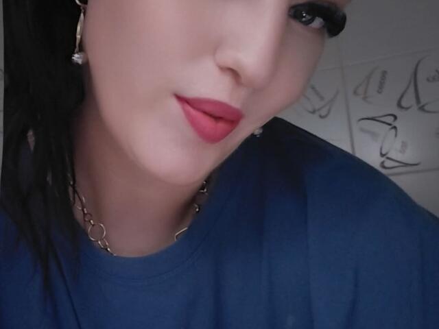 VanessaSquirtt's face with red lips, blue shirt, and wet black hair, wearing a chain necklace and hoop earrings, in a selfie. Her eyes are looking slightly to the side.