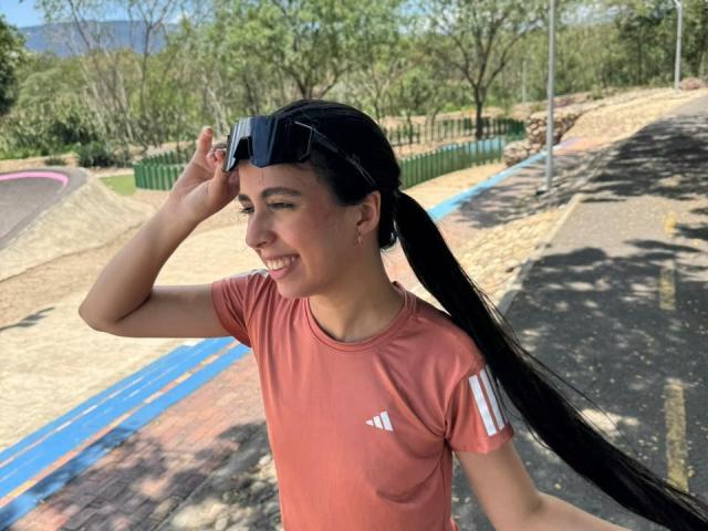 AnnaAlvarez with long black ponytail, wearing a pink Adidas shirt, smiling, adjusting black sunglasses on her head, standing on a sunny outdoor pathway with greenery in the background.