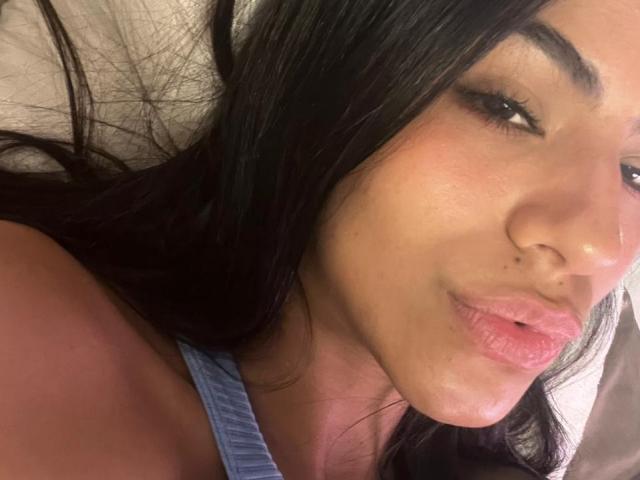 EvaGate ha lunghi capelli neri, un carino piercing al naso e labbra rosa e gonfie, indossa una maglietta blu, appare sexy con un leggero sorriso. Primo piano, illuminazione soffusa..