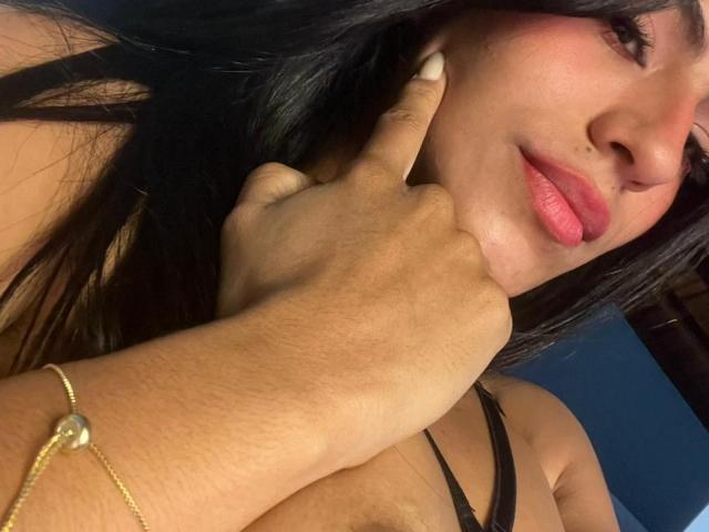 Eva, con i capelli scuri, il rossetto rosa e un sorriso carino, che tocca la guancia con un dito, indossa un top nero con spalline sottili e un braccialetto dorato, mostrando il seno..