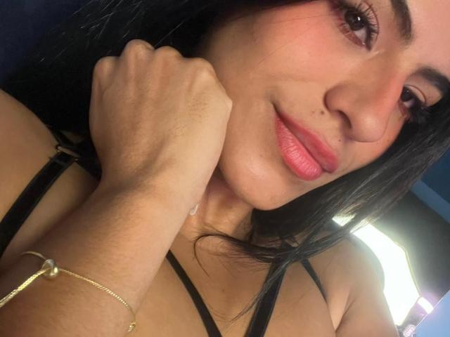 EvaGate with black hair, pink lipstick, and a small mole on her nose, smiling, resting her cheek on her hand, wearing a black strap top, and a gold bracelet.