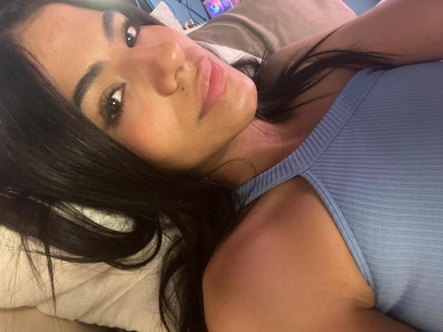 Eva, con lunghi capelli neri, un top a coste blu e labbra piene, è sdraiata, mostrando leggermente il seno e i glutei. Ha un piccolo neo sopra il labbro..